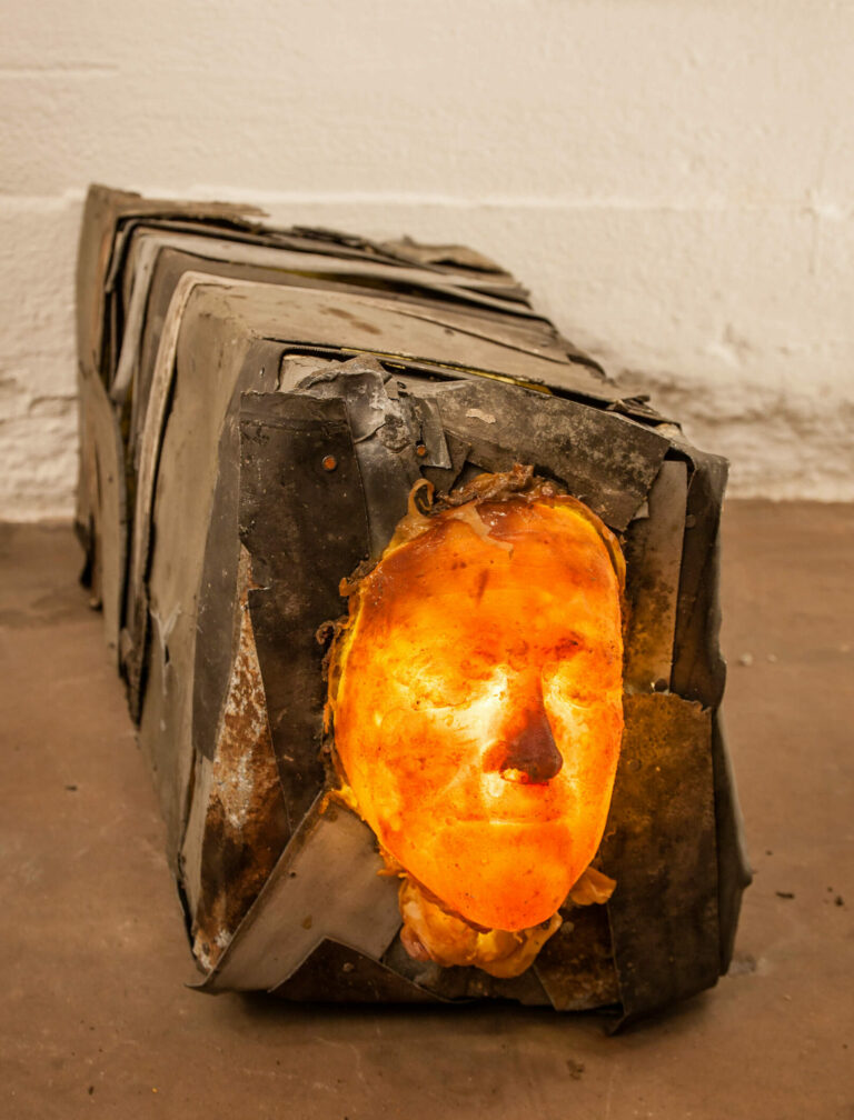 the sculpture shows a mysterious face connected with a body made of metal. illuminated from the inside